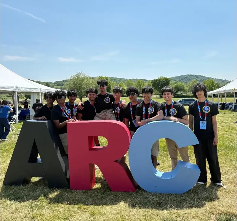 ARC Team B members in black club shirts and lanyards leaning on giant ARC letters at the 2026 American Rocketry Challenge National Finals
