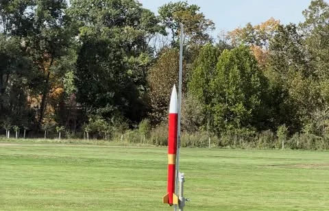 A red and white SBHS student-built competition rocket standing on a launch rail in a grass field