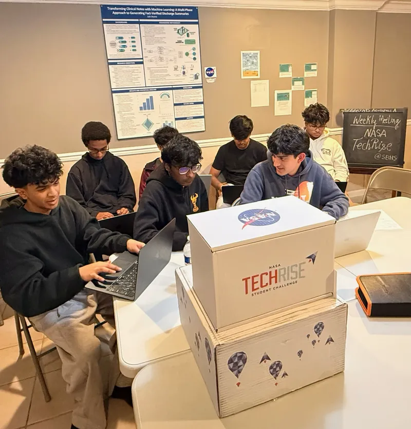 Team members working on laptops around the NASA TechRise kit boxes at a weekly meeting